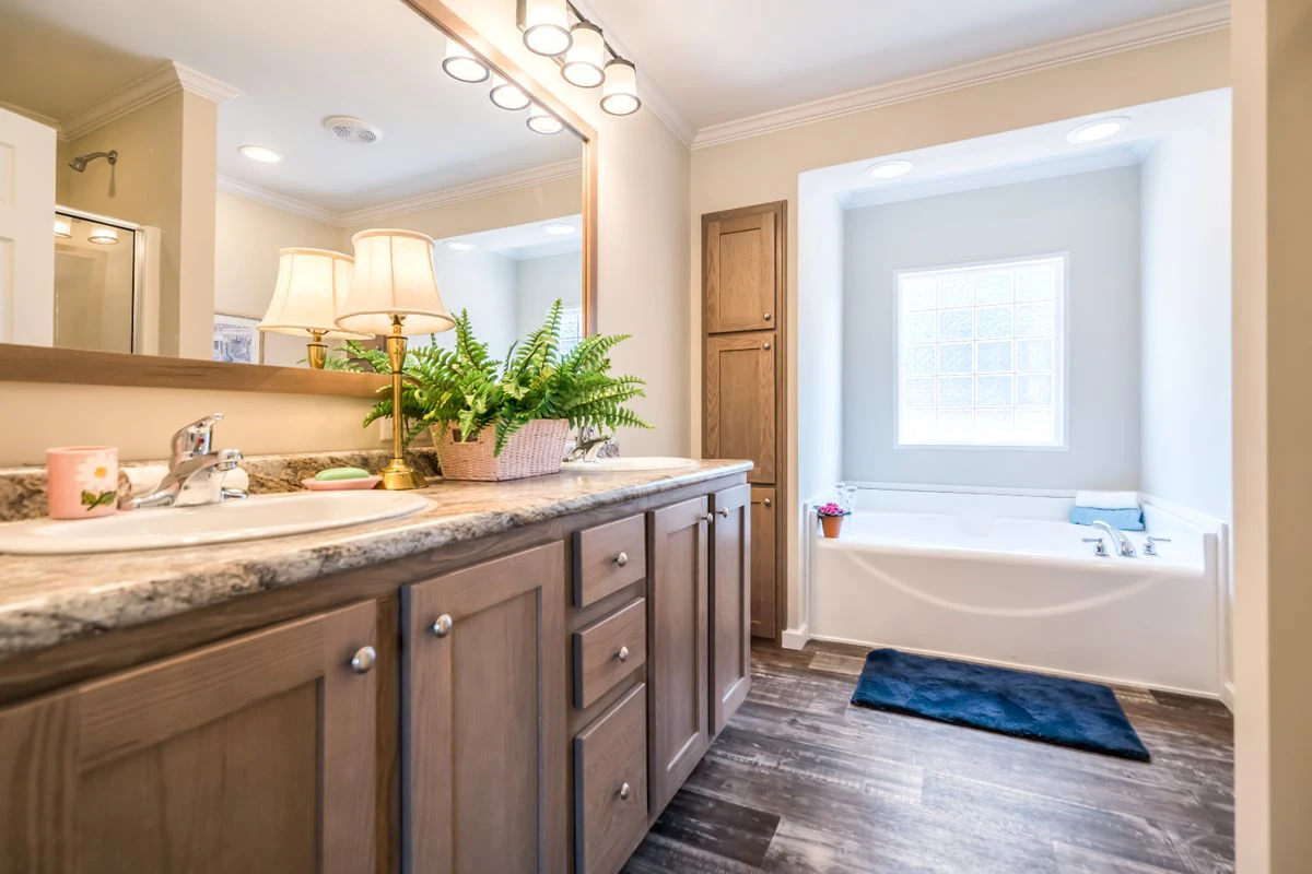 Spacious bathroom with a double vanity, large mirror, potted plant, wood cabinets, a soaking tub by a window, and dark wood flooring.
