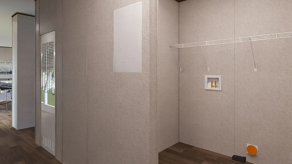 Empty laundry room with beige walls, wire shelf, utility connections, and a recessed utility box; adjacent room with visible flooring and partial kitchen appliances.