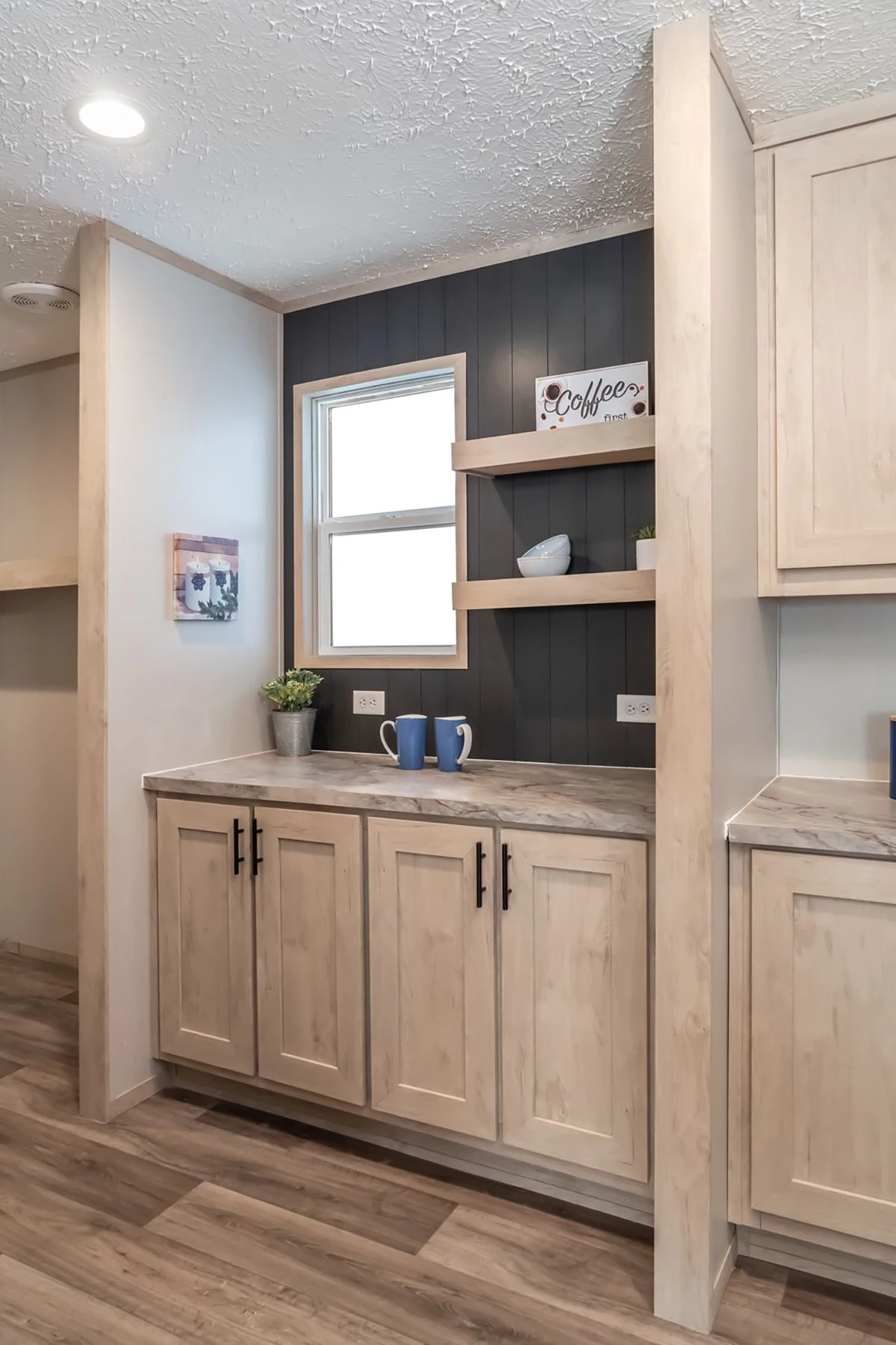 A kitchen area with light wood cabinets, a window, open shelves holding decor items, a small plant, two mugs, and a "coffee" sign on a dark accent wall.