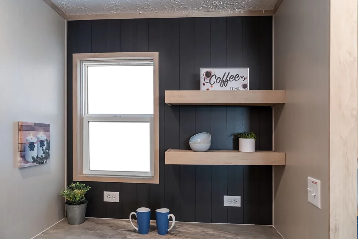 A small kitchen nook with two blue mugs on the counter, floating shelves holding a bowl, a plant, and a “Coffee first” sign, and a window providing natural light.