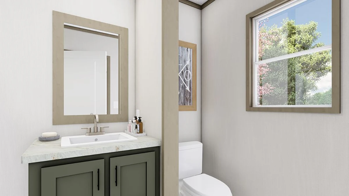Modern bathroom with a green vanity, sink, and mirror on the left, a toilet in the middle, framed artwork, and a window showing trees on the right.