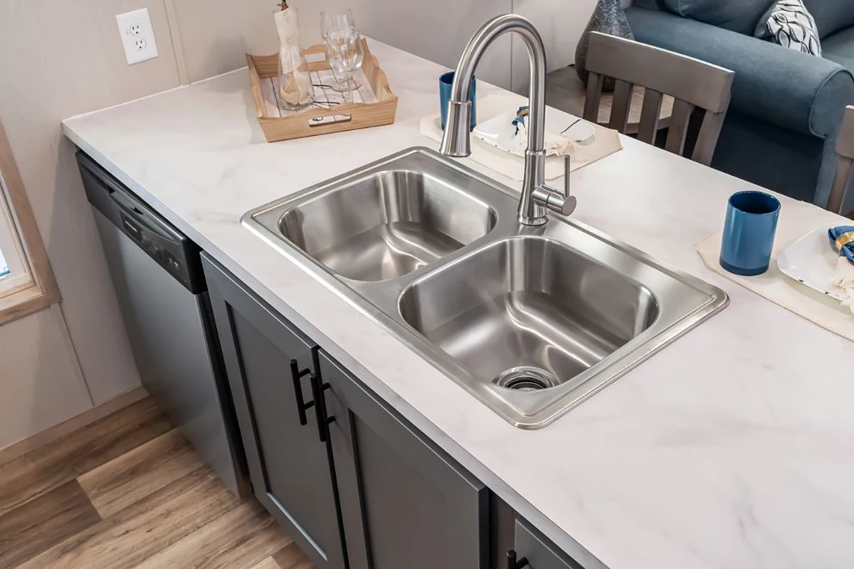 A modern kitchen features a double-basin stainless steel sink with a high-arc faucet, dark cabinetry, and a marble countertop. A dishwasher and tray with glasses are nearby.