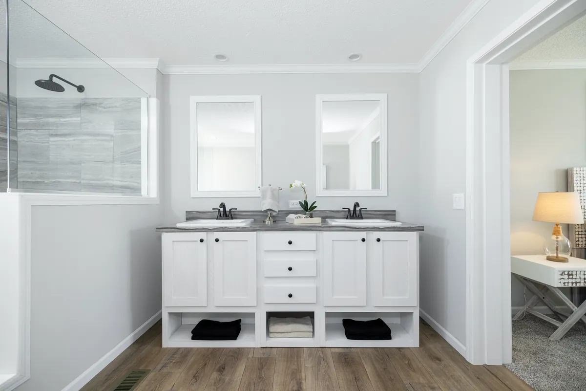 b0676c0e-dccd-40ef-a201-793ab5158b76 Modern bathroom with double sinks, two mirrors, white cabinets, gray countertops, an orchid, and a glass shower on the left; adjacent bedroom visible on the right.