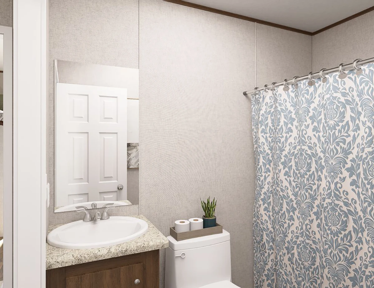 A bathroom with a sink, mirror, and toilet; a small plant and toilet paper sit on the toilet tank, and a shower curtain with a blue pattern hangs beside them.