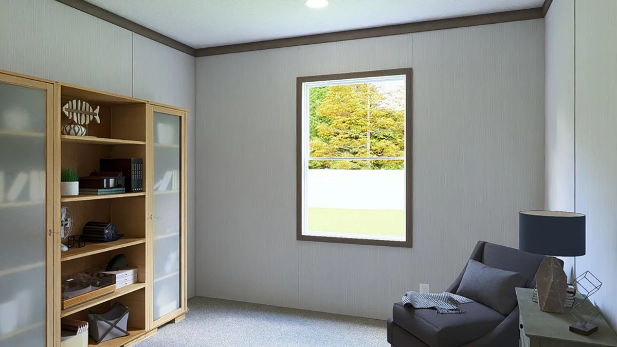 A small room with a window, a gray armchair, a side table, and a wooden bookshelf against white walls. Green trees are visible outside the window.