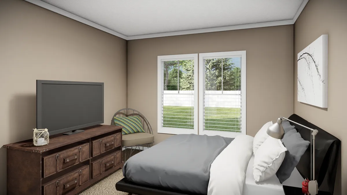 A bedroom with a bed, dresser with a TV, chair in the corner, wall art, and two windows showing a view of a green yard with trees.