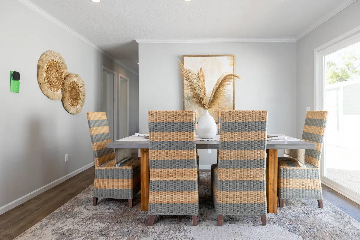 bebdc64c-2172-4714-a9bc-467ed2905a77 A dining room features a wooden table with six wicker chairs, a large vase with pampas grass centerpiece, wall decor, and natural light from a sliding glass door.