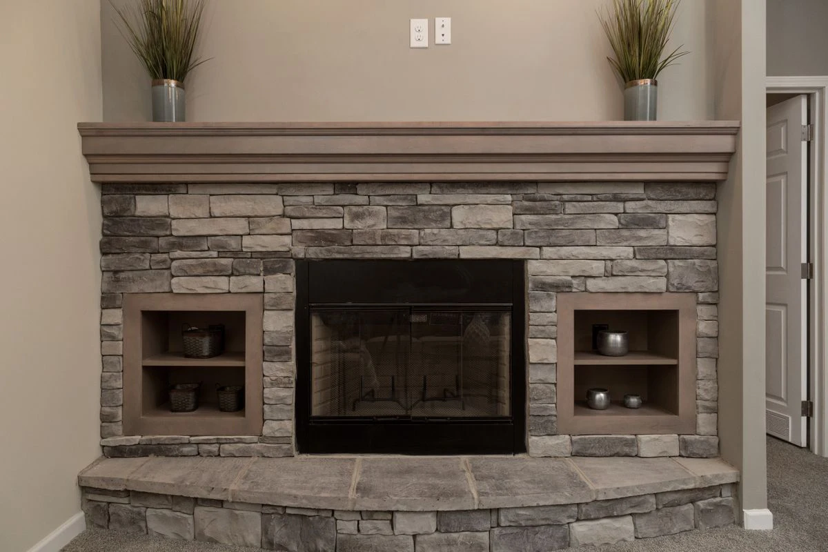 Stone fireplace with a wooden mantel, two built-in shelves holding decorative items, and two potted plants on top; electrical outlets above the mantel.