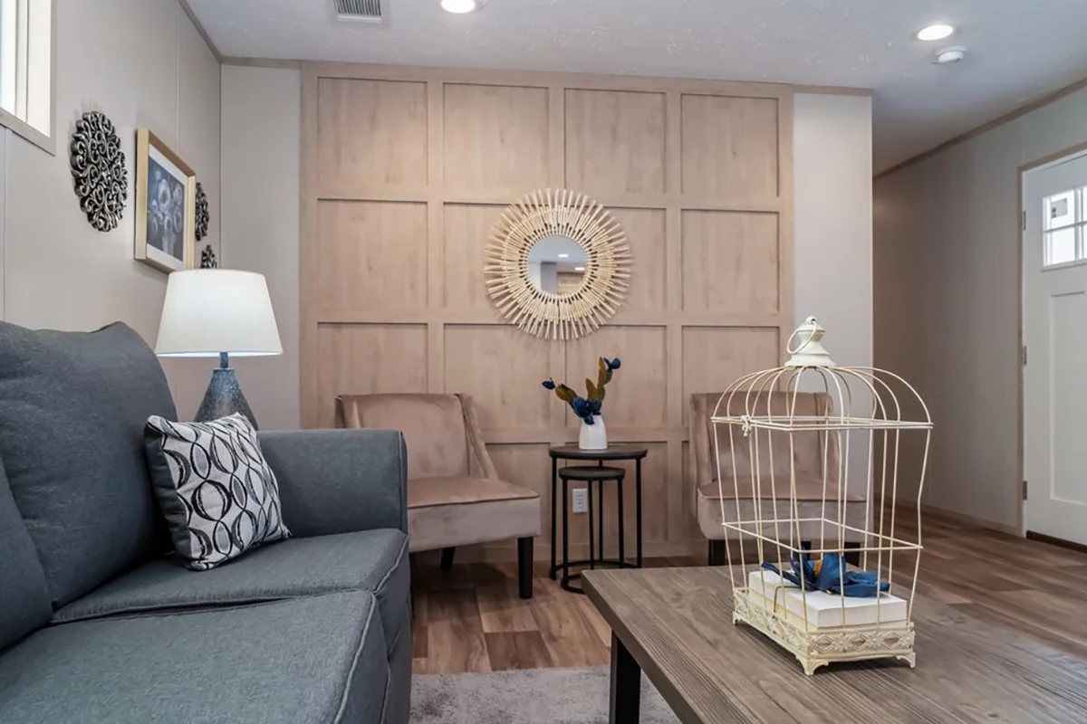A living room with a gray sofa, two beige chairs, a wooden coffee table with a birdcage decor, and a wood-paneled accent wall with a round mirror.