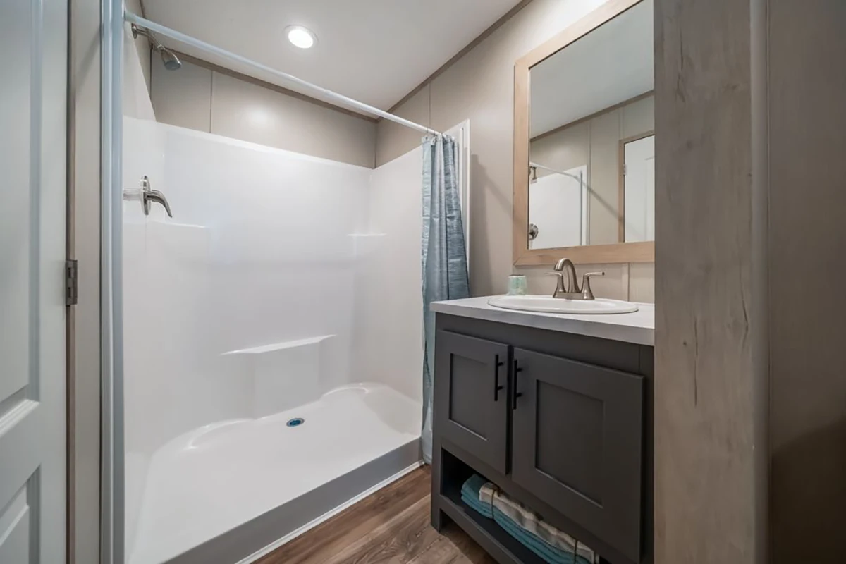 Bathroom with a white shower stall, a gray vanity with sink and mirror, a blue shower curtain, and wood-look flooring. Towels are stored on a shelf under the sink.