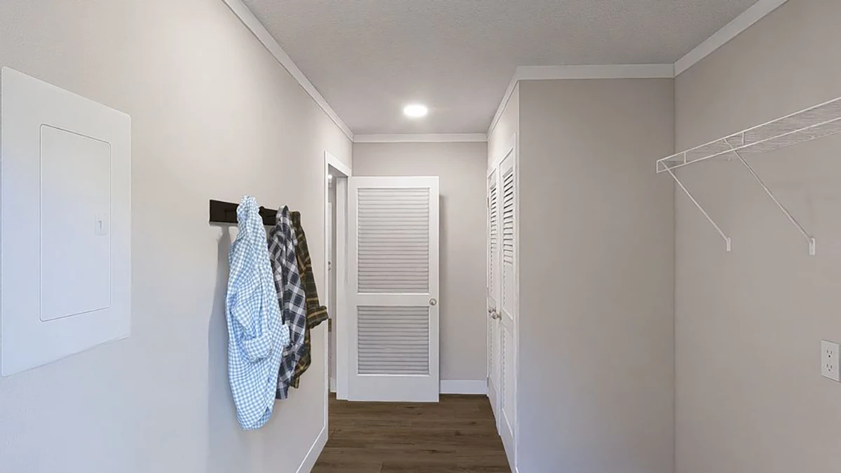 A hallway with beige walls, a wooden floor, wall hooks holding shirts, a wire shelf, and doors at the end and right side.