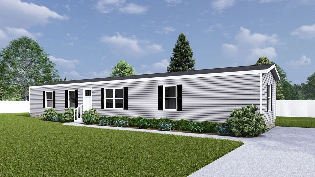 Single-story gray manufactured home with black shutters, white trim, small entry steps, landscaped shrubs, and a driveway, set against a grassy yard and trees under a partly cloudy sky.