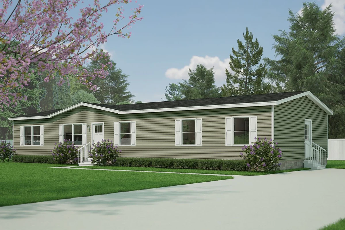 d05336aa-5886-434a-8e99-146e559f4041 A green manufactured home with white trim, multiple windows, two small porches with railings, set on a grassy lawn with trees and a paved driveway.