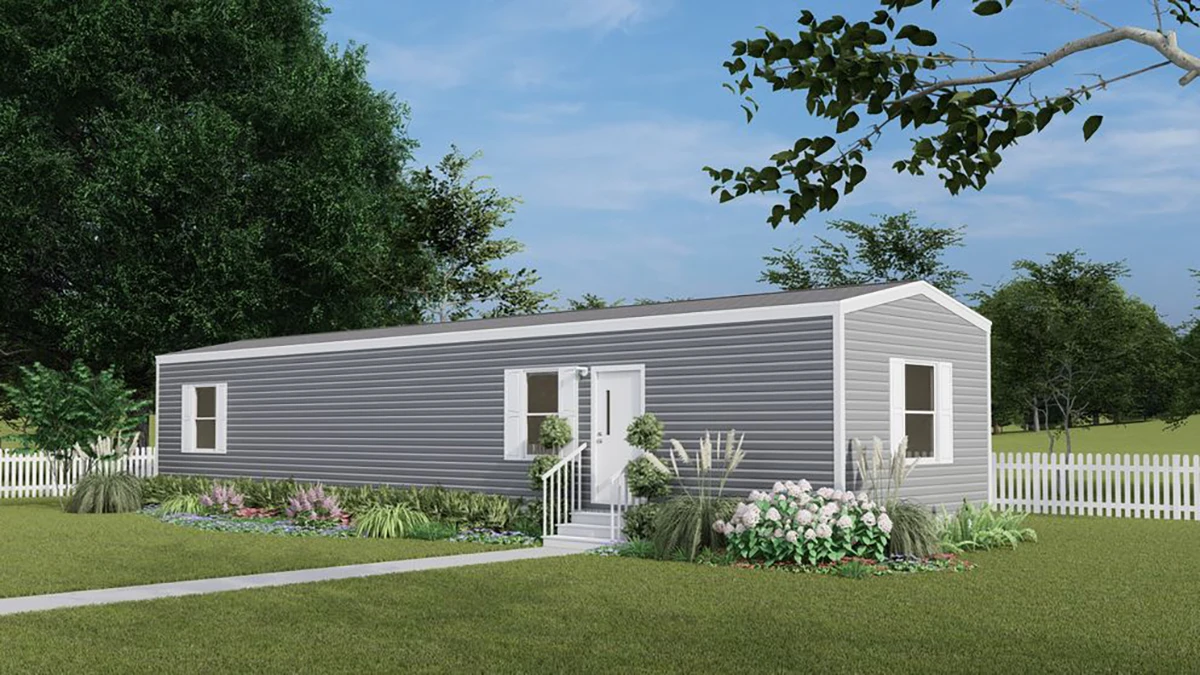 A gray single-wide manufactured home with white trim, small front steps, landscaped garden, white picket fence, and green lawn under a blue sky.