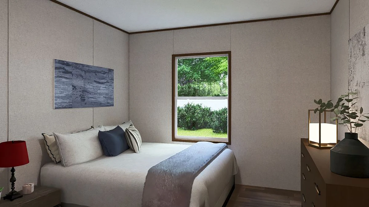 A small bedroom with a bed, neutral bedding, a nightstand with a red lamp, a dresser with decor, a wall painting, and a window showing greenery outside.