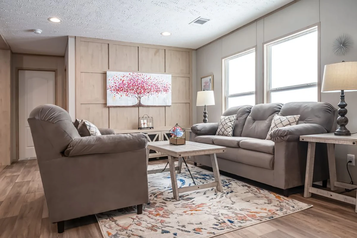 A living room with two gray sofas, a wooden coffee table, two side tables with lamps, a patterned rug, and a wall art piece featuring a red tree. Natural light comes through two windows.