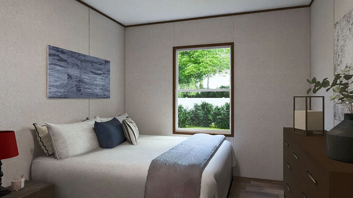 A small bedroom with a neatly made bed, two pillows, a red lamp on a side table, a dresser, and a window looking out onto greenery.