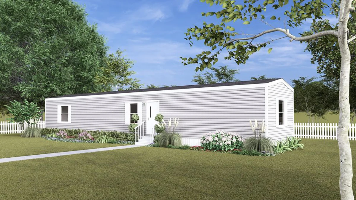 A white, single-wide mobile home with a small front garden and a white picket fence, set on a grassy lawn with trees in the background.