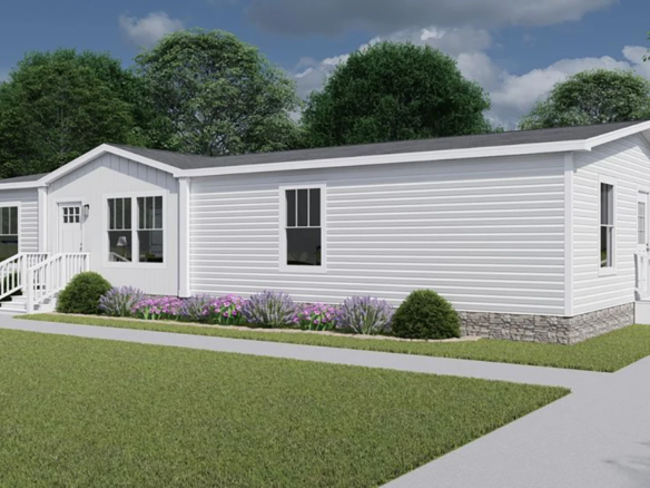 A white manufactured home with a gable roof, steps leading to front and back doors, flower beds along the side, and surrounded by grass and trees under a partly cloudy sky.