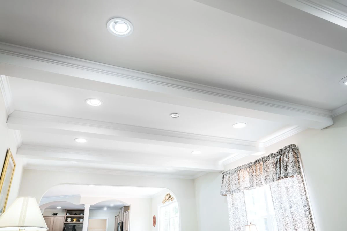 White coffered ceiling with recessed lighting in a bright living space, featuring crown molding and a window with patterned curtains.