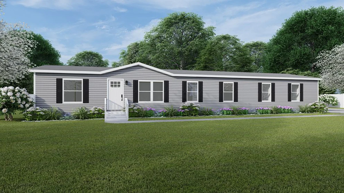 A single-story, gray manufactured home with black shutters and a small porch, surrounded by green lawn and trees under a blue sky.