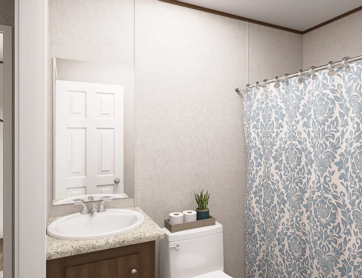 A bathroom with a sink and mirror, a toilet holding two toilet paper rolls and a small potted plant, and a shower with a blue and white patterned curtain.