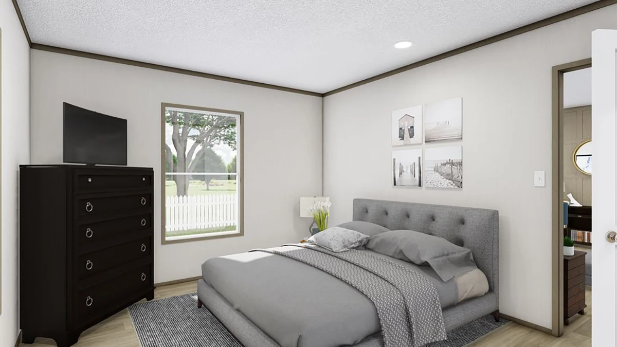 A modern bedroom with a gray upholstered bed, gray bedding, a dark dresser with a TV, wall art, and a window showing a yard with trees and a white fence.