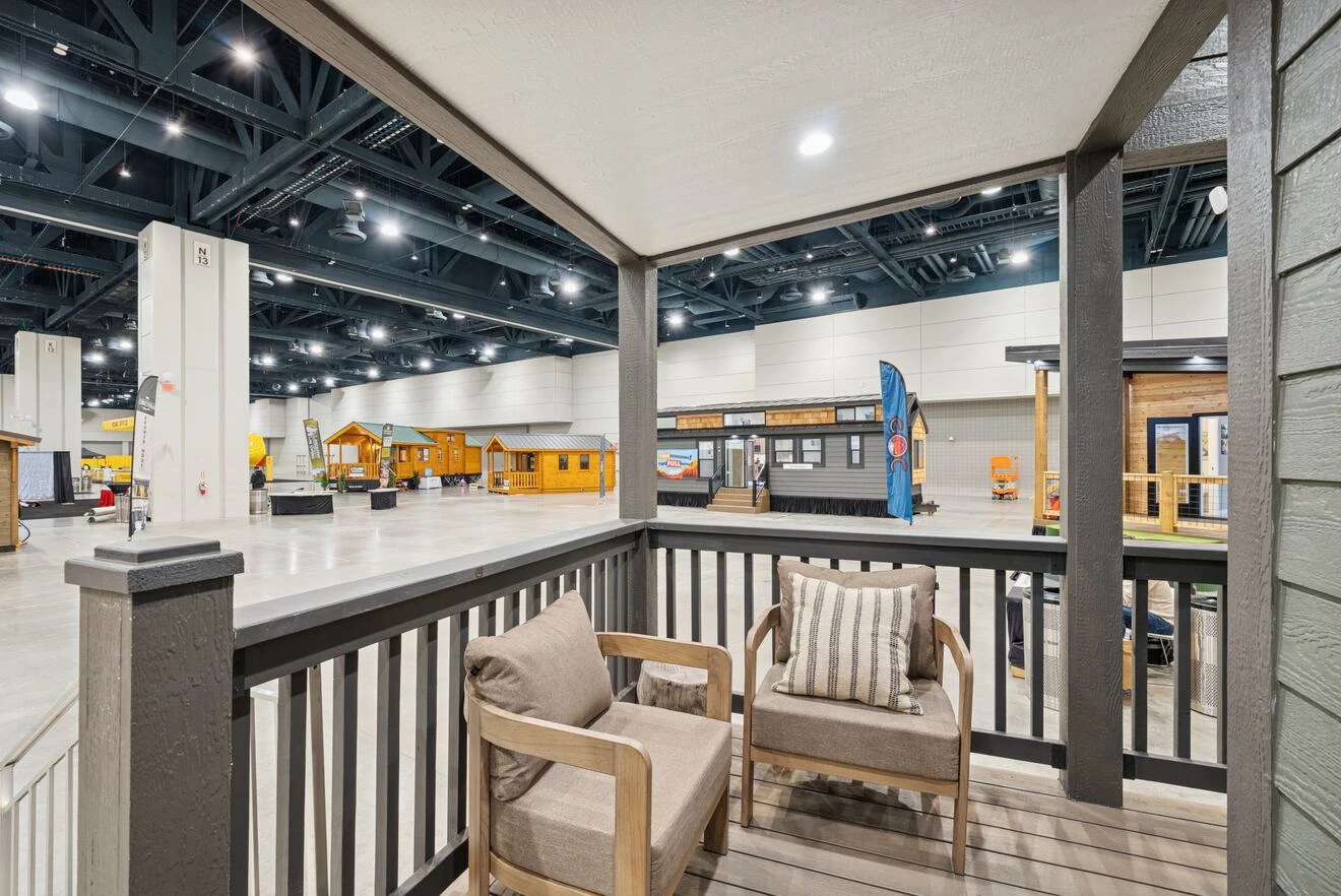 View from a porch with two cushioned chairs, overlooking a spacious indoor exhibition hall featuring several small house displays.