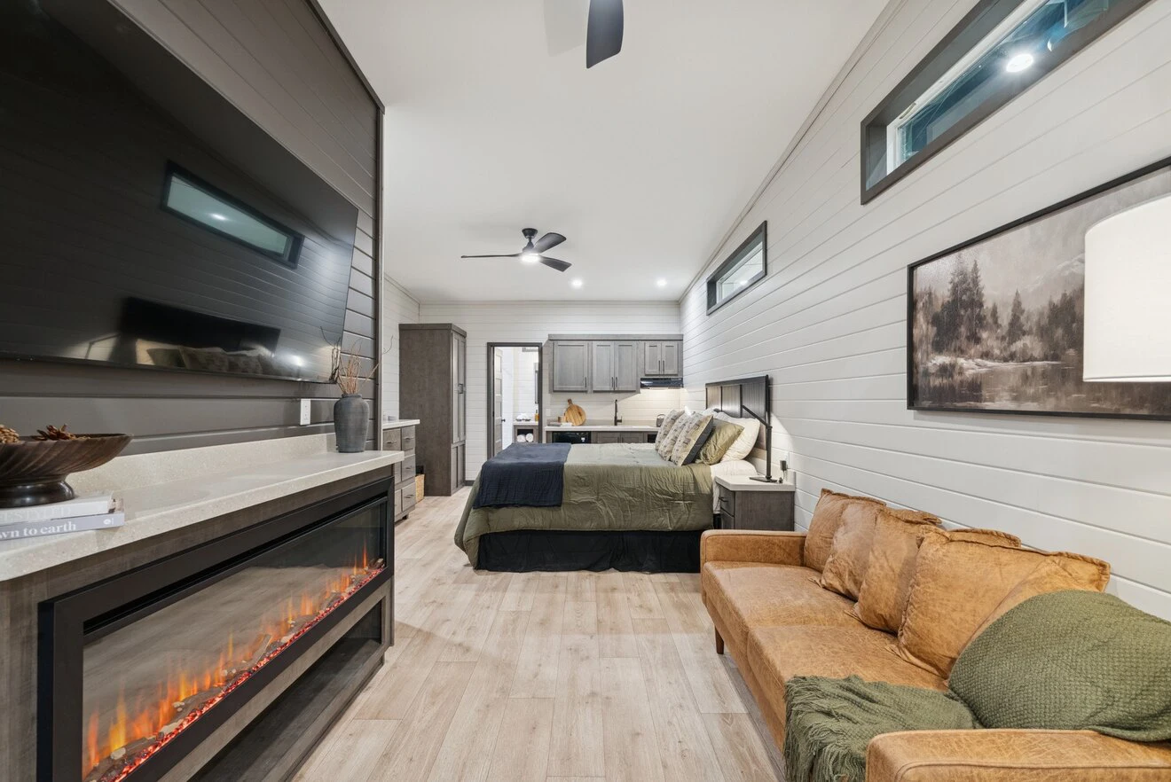 Modern bedroom with a wall-mounted TV above an electric fireplace, brown leather sofa, bed with green and black bedding, wood floors, and neutral decor.