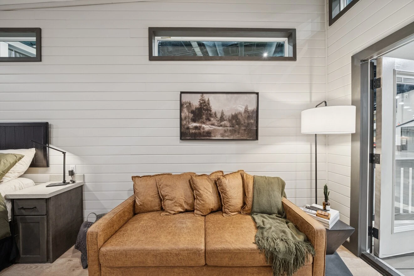A brown leather sofa with three cushions sits against a white shiplap wall, beneath a landscape painting. A floor lamp and side table with a plant are on the right.
