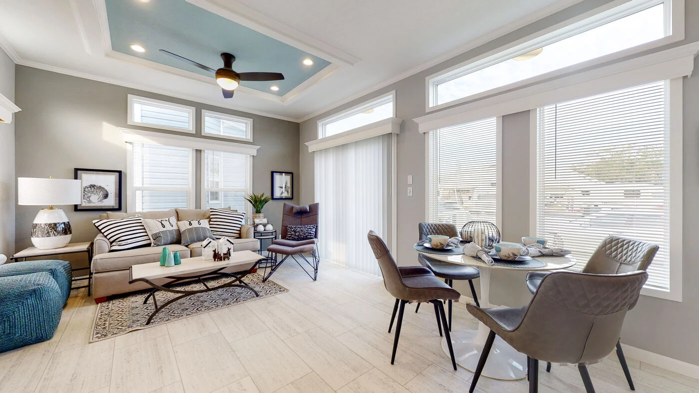 Modern living and dining area with large windows, light wood flooring, a gray sofa, round dining table with four chairs, and decorative ceiling with fan.