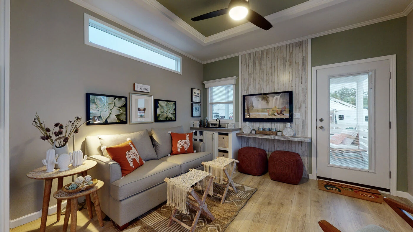 A cozy living room with a gray sofa, orange pillows, wall art, a mounted TV, two red poufs, and a door leading outside. Light wood floor and neutral walls with decorative accents.