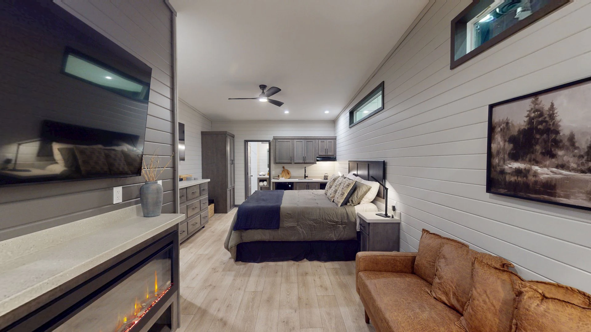 Modern bedroom with wood flooring, a large bed, gray cabinetry, wall-mounted TV, electric fireplace, brown sofa, and landscape artwork on a white shiplap wall.
