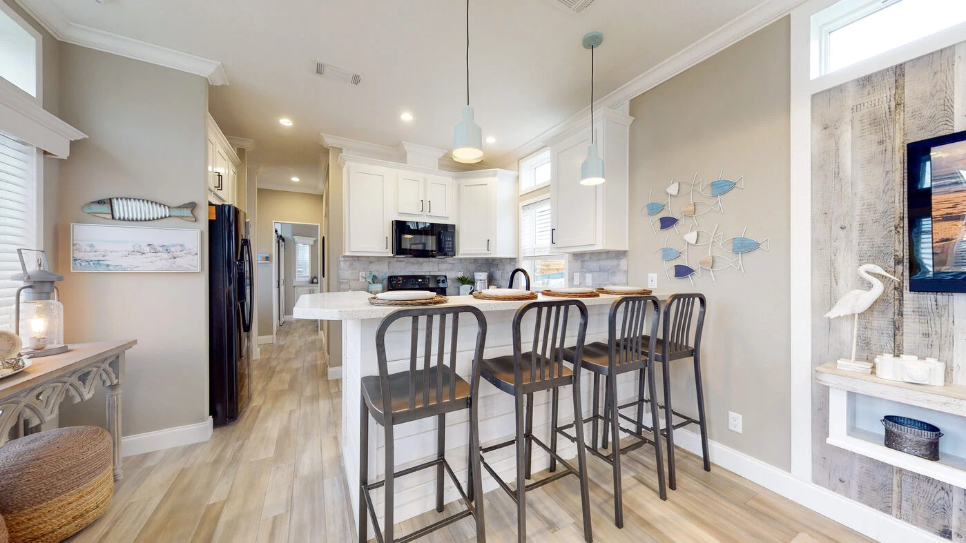 Modern kitchen with a white island, three bar stools, black appliances, light wood flooring, and coastal-themed decor including fish wall art and a heron sculpture.