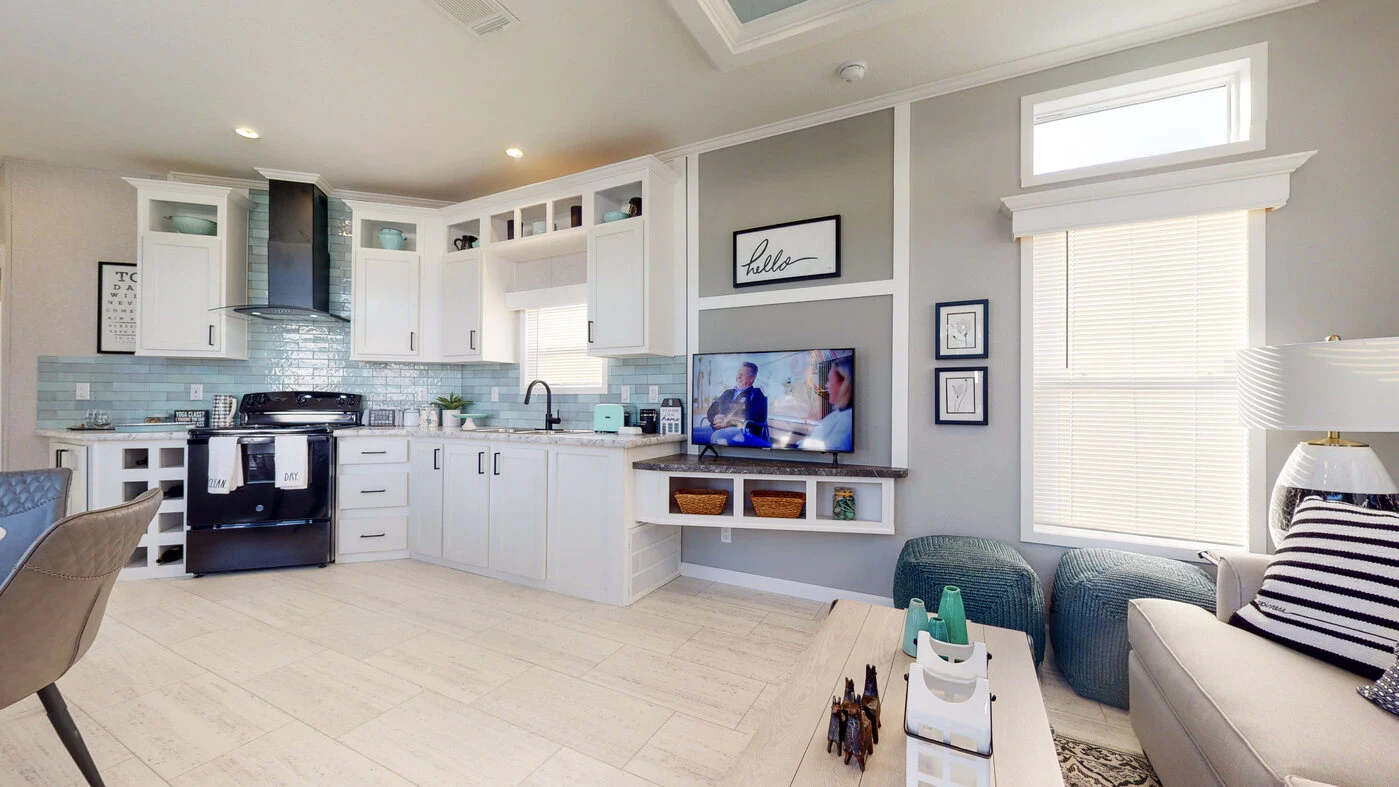 Modern living room and kitchen with white cabinets, light wood flooring, wall-mounted TV, and blue accents. Large window with blinds lets in natural light.