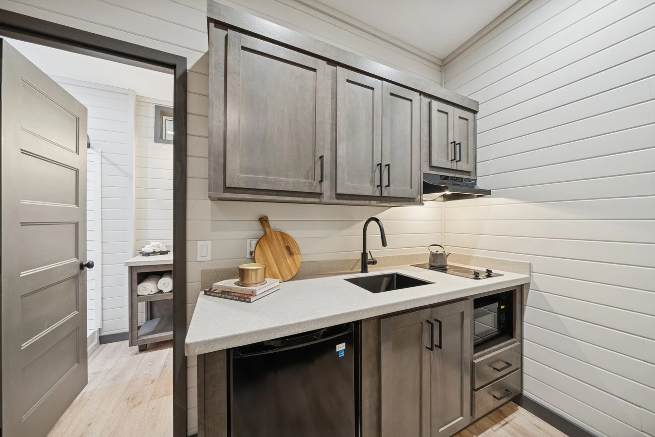 Compact kitchen with gray cabinets, a small sink, stove, microwave, mini-fridge, and cutting boards on the counter; door leads to a bathroom with towels on open shelves.
