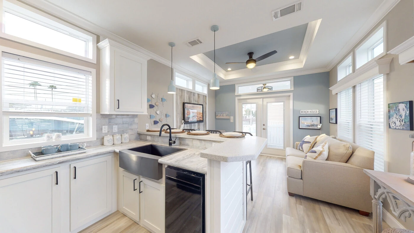 Modern kitchen and living area with white cabinets, farmhouse sink, island, light wood flooring, and a small sofa against a blue accent wall. Large windows provide ample natural light.