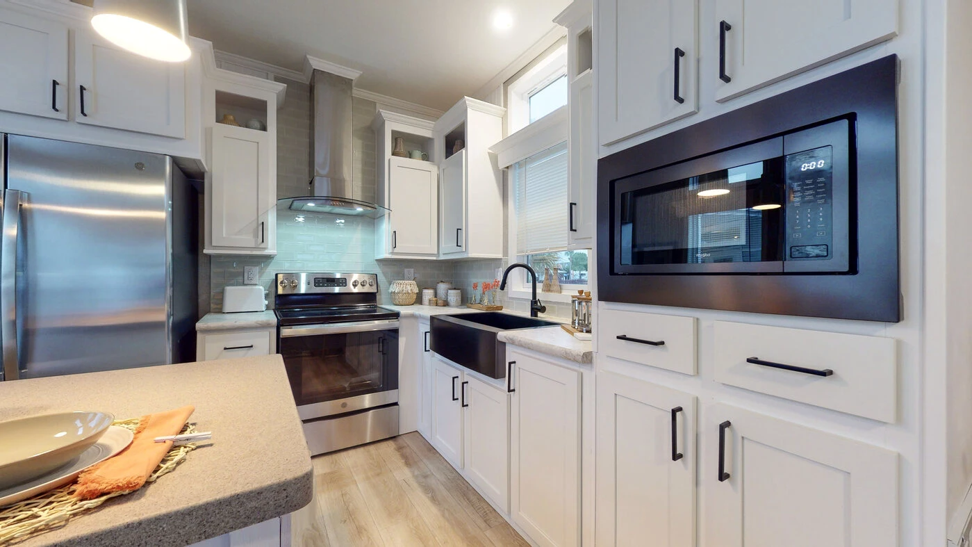 Modern kitchen with white cabinets, stainless steel appliances, a black farmhouse sink, built-in microwave, and a light wood floor. Countertops hold various kitchen items and decor.