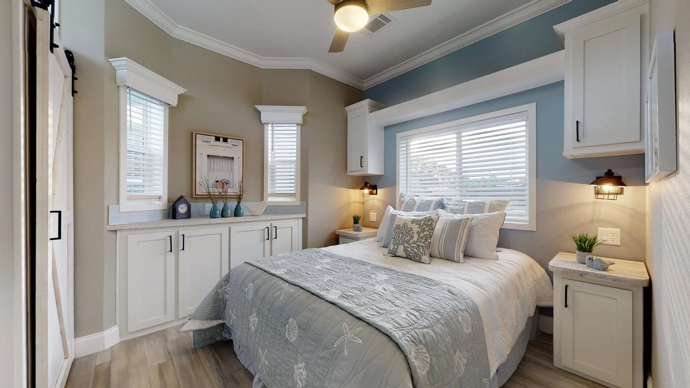 A tidy bedroom with a double bed, light blue and white bedding, built-in cabinets, two windows with blinds, and a ceiling fan.