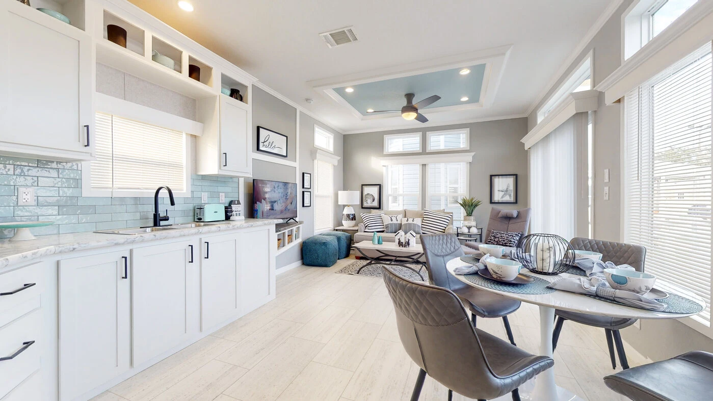 Modern open-plan kitchen and living area with white cabinetry, dining table set for four, teal accents, and large windows allowing natural light.