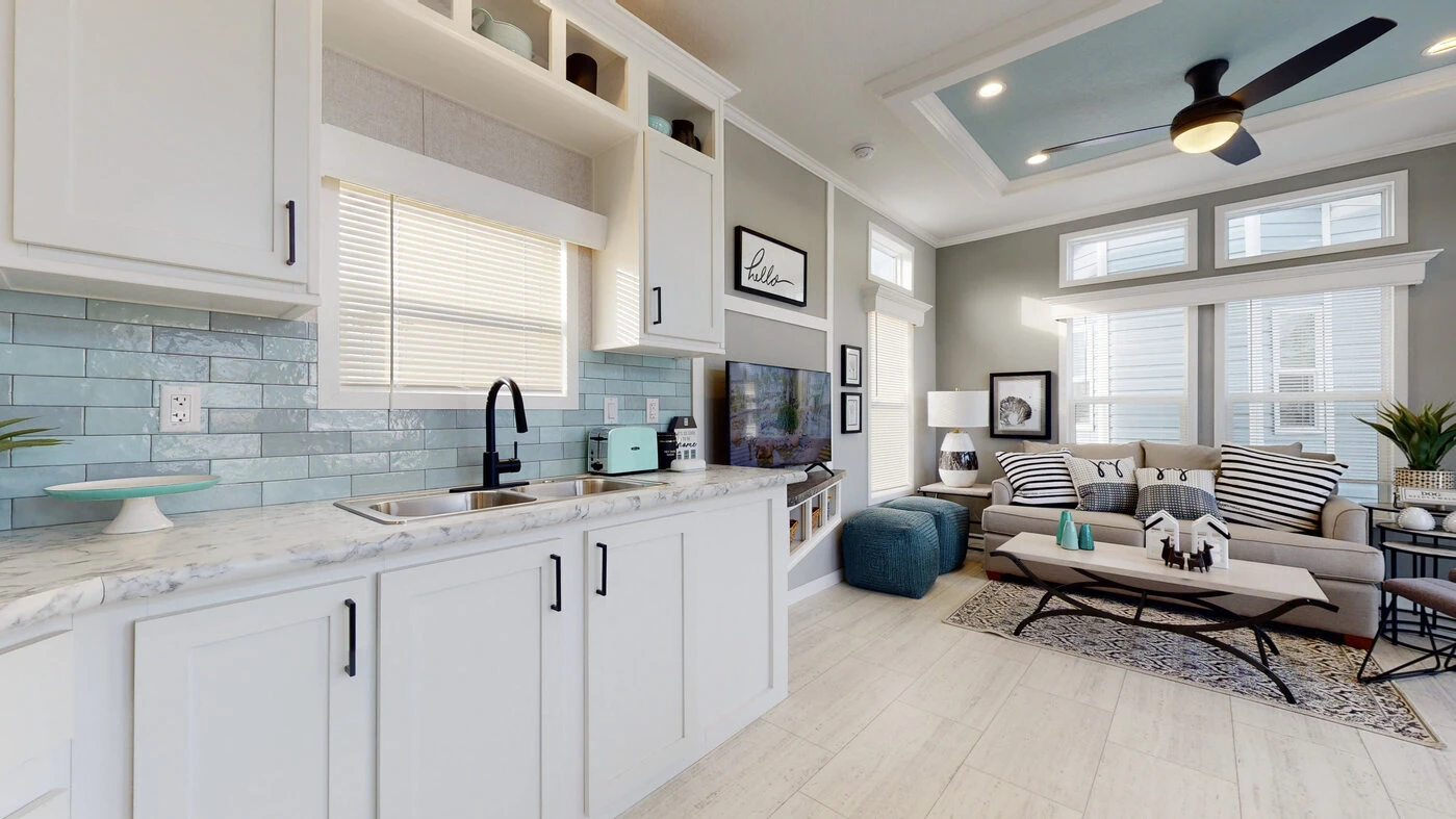 Modern open-plan kitchen and living room with white cabinets, light blue tiled backsplash, marble countertop, grey sofa, coffee table, and ceiling fan. Natural light streams through windows.