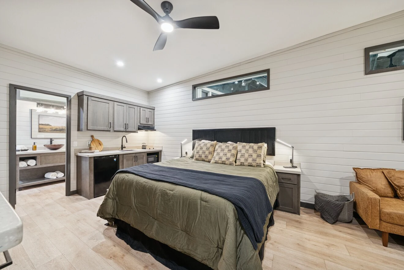 Modern bedroom with a large bed, kitchenette, ceiling fan, and ensuite bathroom; neutral tones and wood paneling create a cozy atmosphere.