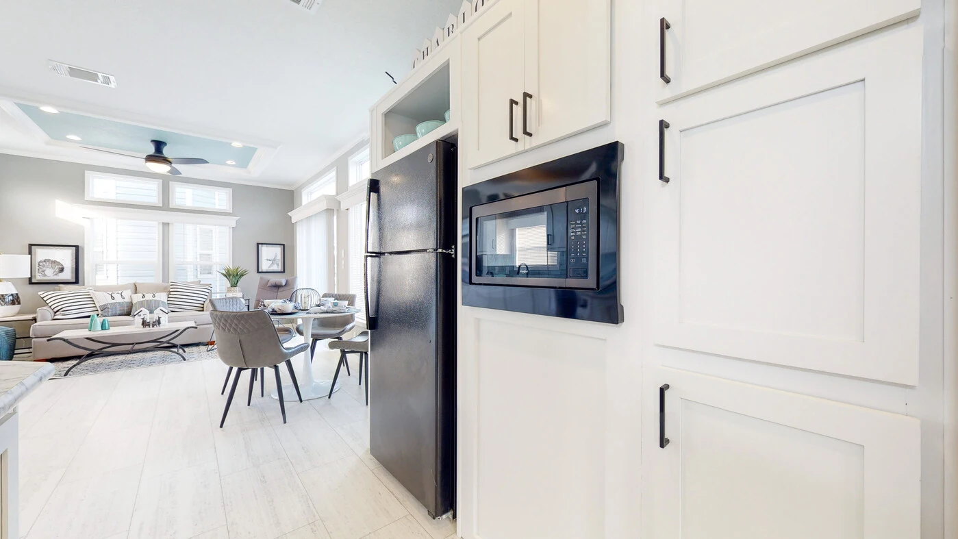 Modern kitchen with white cabinets, a built-in microwave, black refrigerator, and an adjacent dining and living area with large windows and neutral decor.