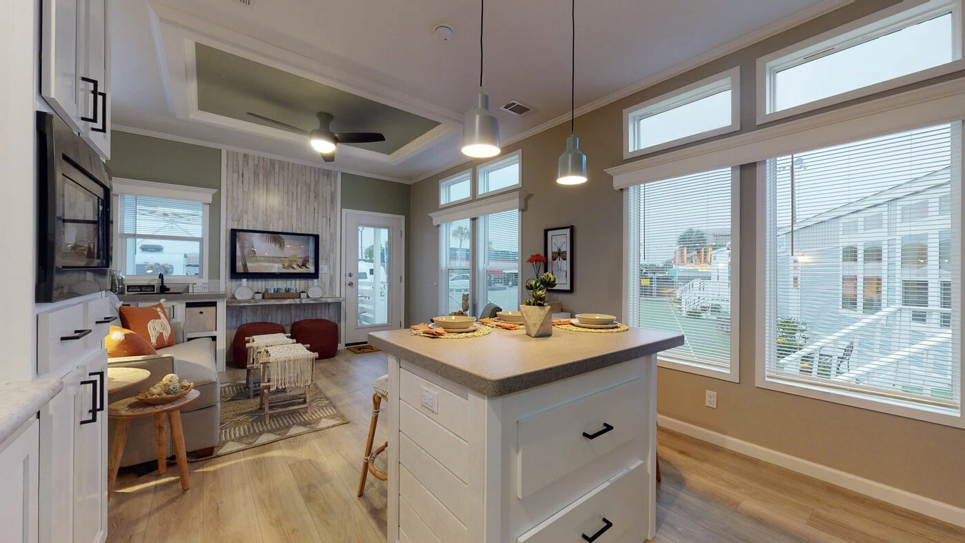Modern kitchen and living area with large windows, island with drawers, pendant lights, and a cozy seating area. Daylight streams in, highlighting a clean, open layout.