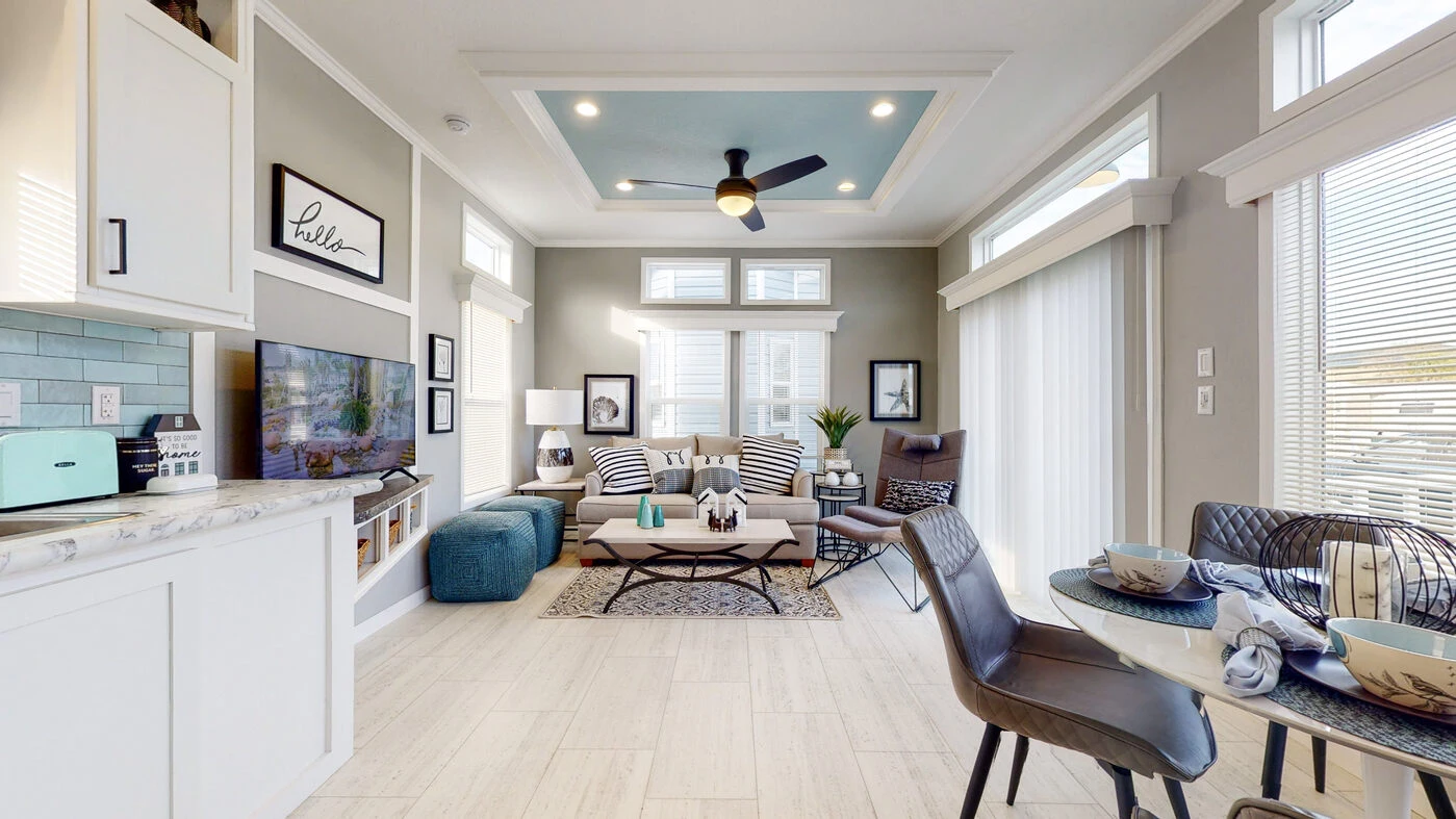 Modern living room with a decorative ceiling, neutral walls, striped sofa, armchair, TV, and adjacent dining area set with dishes. Large windows and sliding glass doors let in natural light.