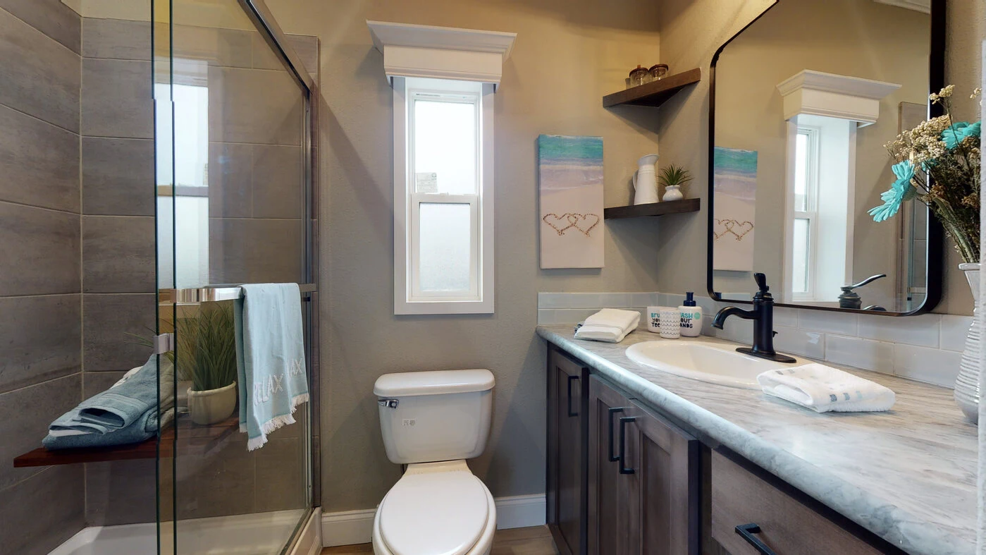 Modern bathroom with glass shower, toilet, large mirror, sink with toiletries, wood cabinets, and wall art next to a window.