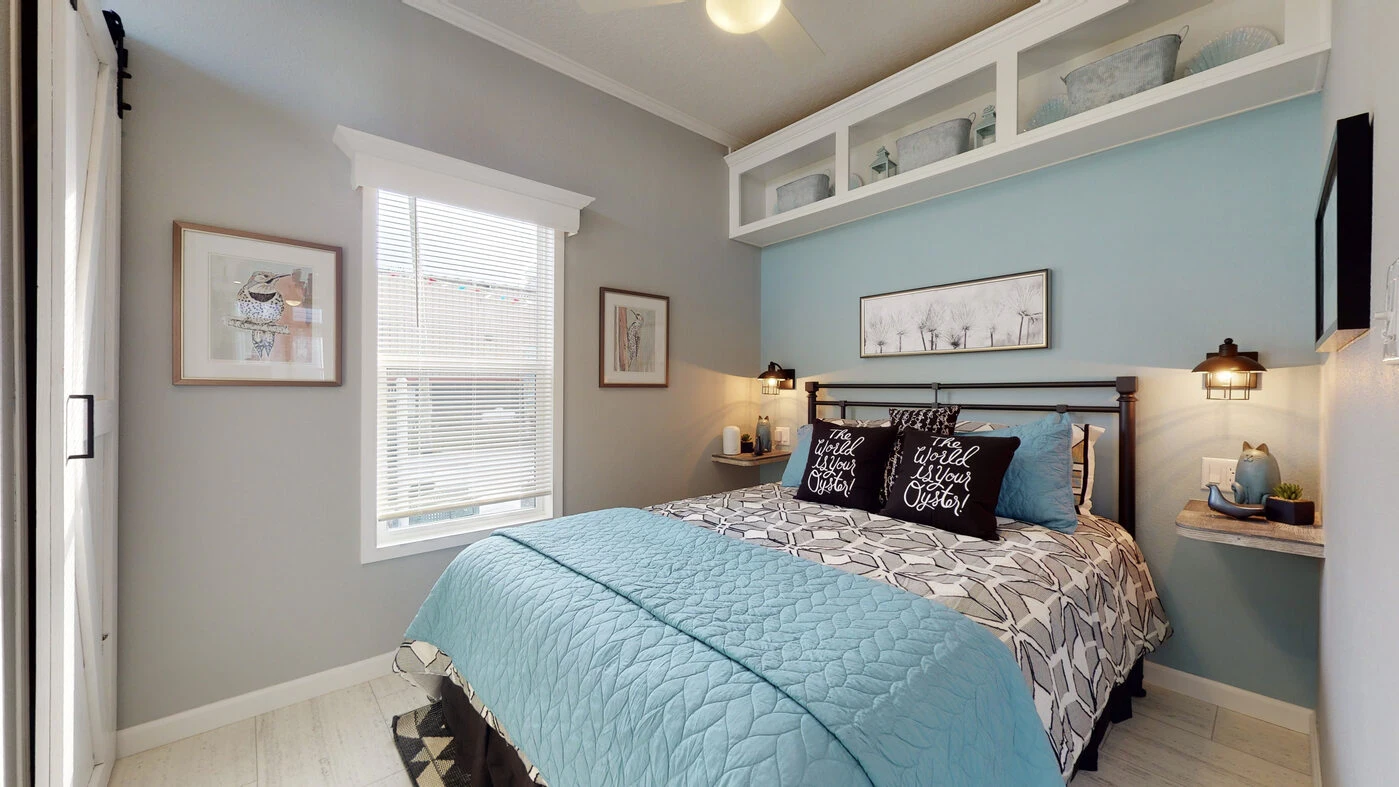 A small bedroom with a blue and gray theme, queen bed, wall art, built-in shelves, two bedside lamps, and a window letting in natural light.