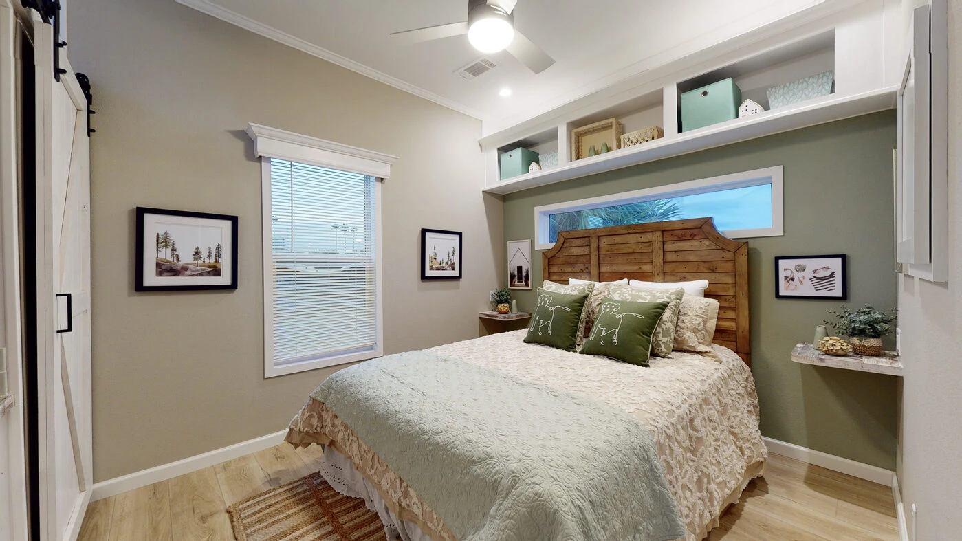 A cozy bedroom with a wooden headboard, green and beige bedding, shelves above the bed, framed art on the walls, and a window with blinds letting in natural light.