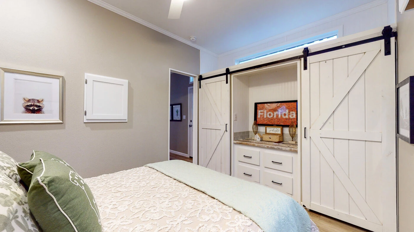 Bedroom with a made bed, beige walls, framed artwork, and a white built-in closet with sliding barn doors and a television inside.
