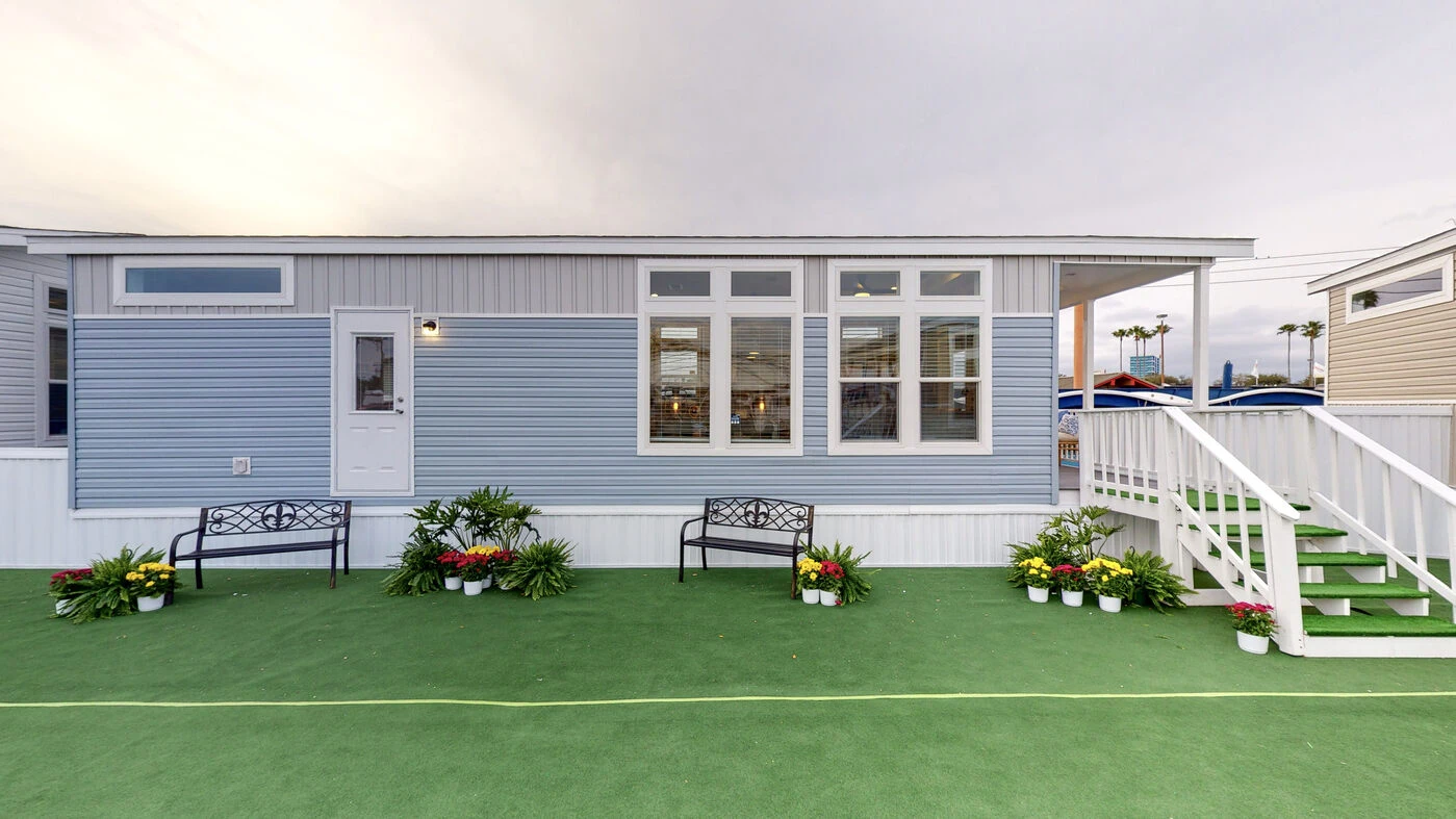 A light blue manufactured home with white trim, a small porch with stairs, two benches, and potted plants on a green artificial turf surface.